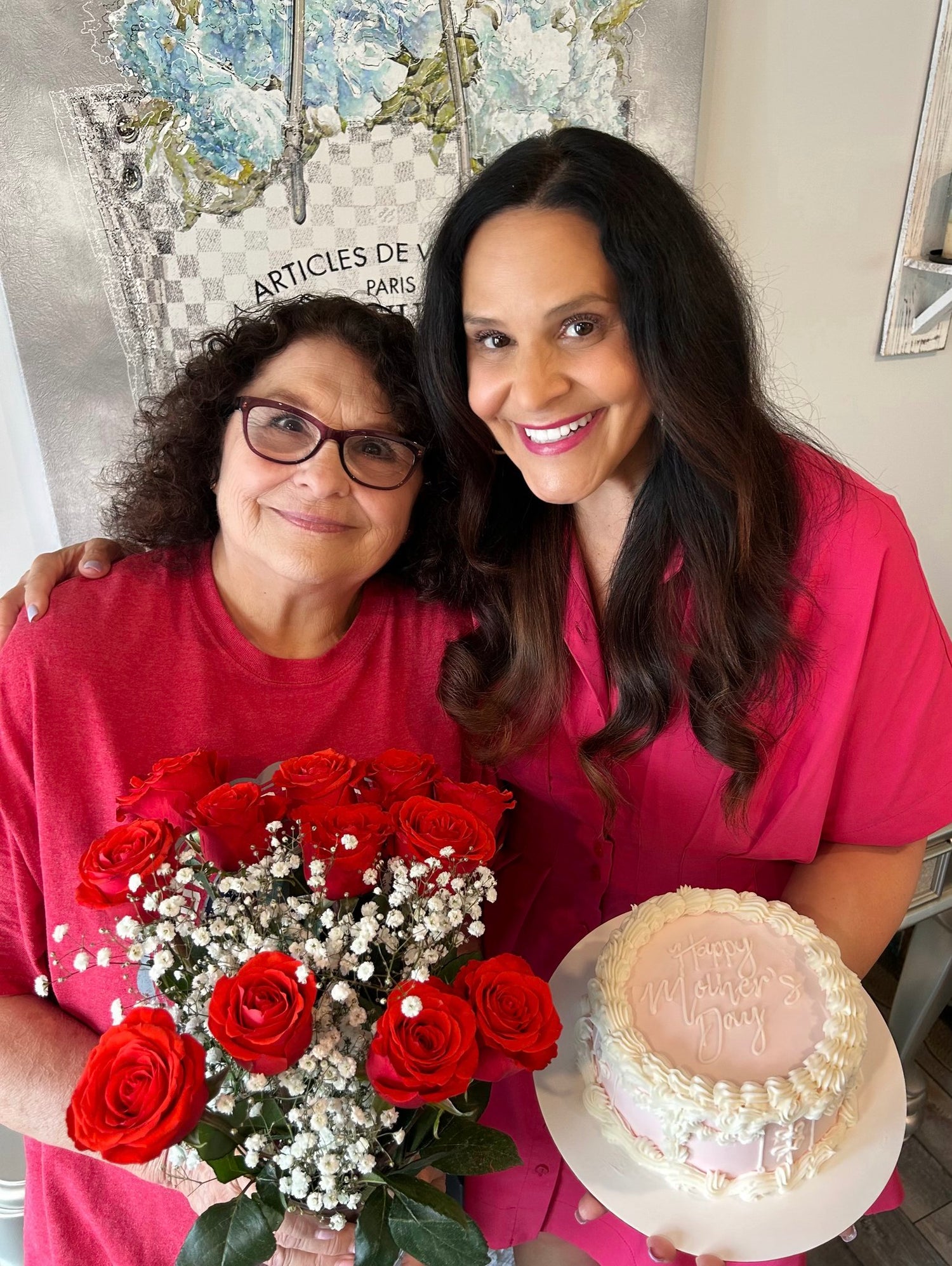 Two women in red shirts holding a bouquet of red roses and a cake with 'Happy Mother's Day' text