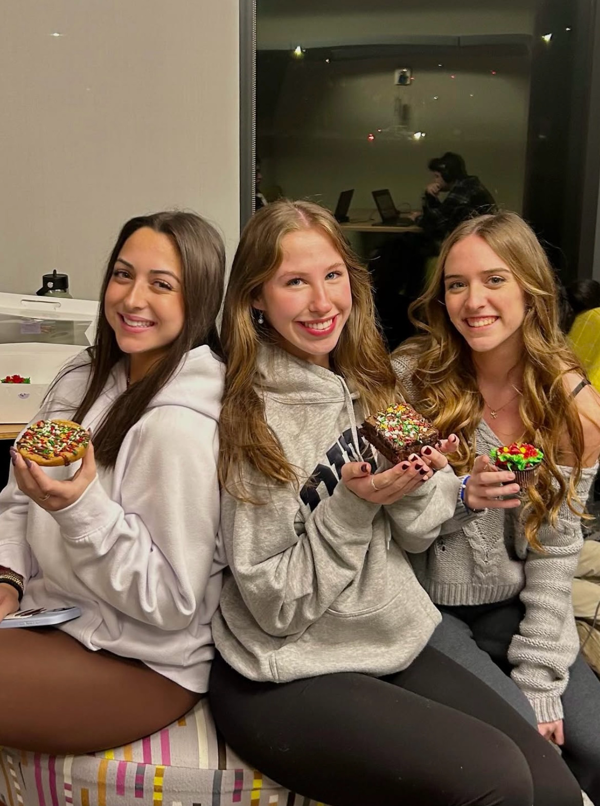 Three women sitting together holding a cookie, brownie, and cupcake in a dorm study area.