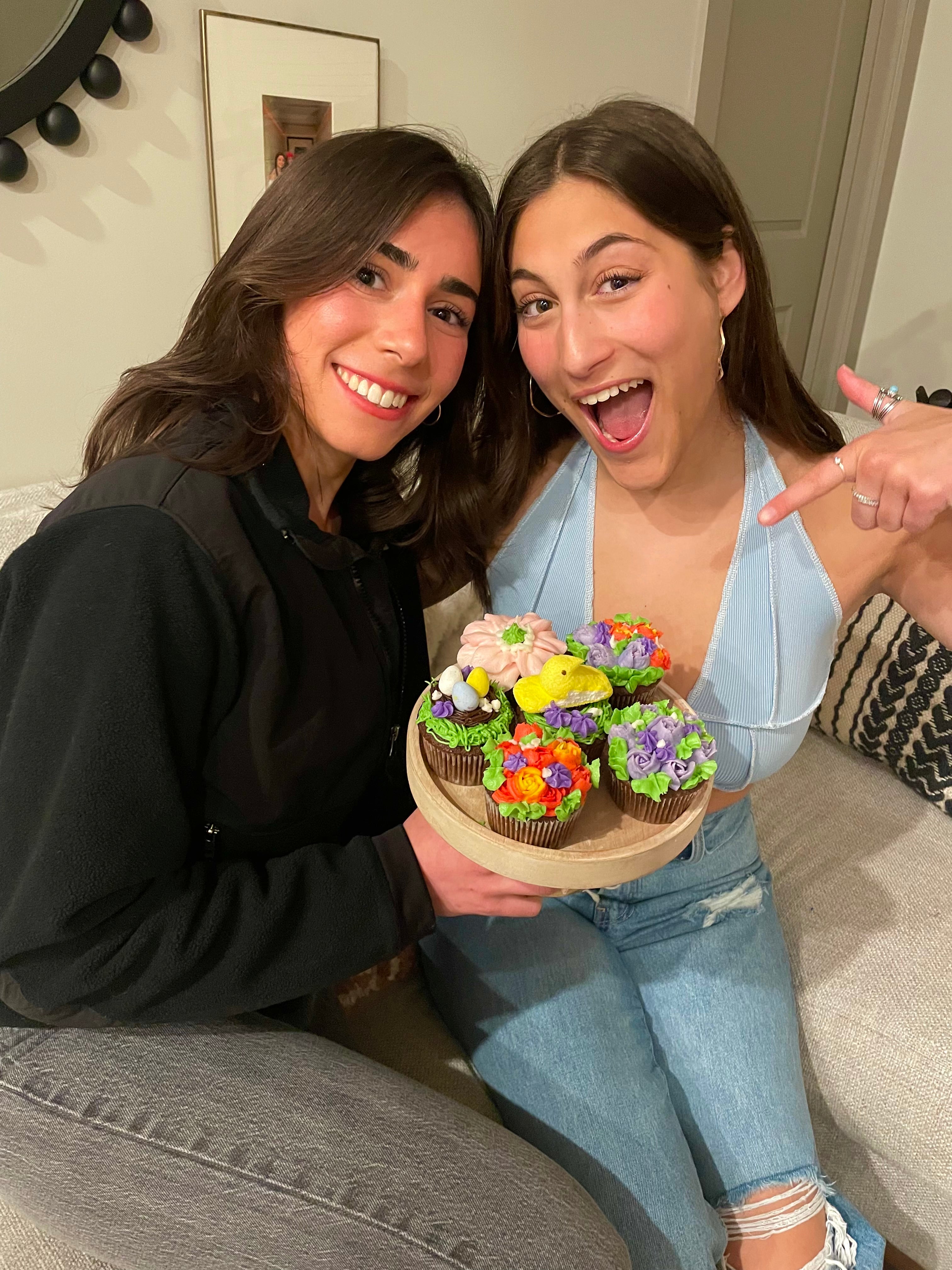 Two women holding a tray of decorated cupcakes.