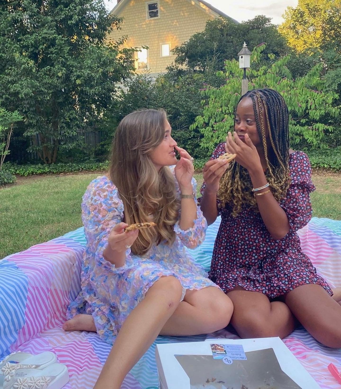 Two women sitting on a colorful blanket outdoors, eating cookies.