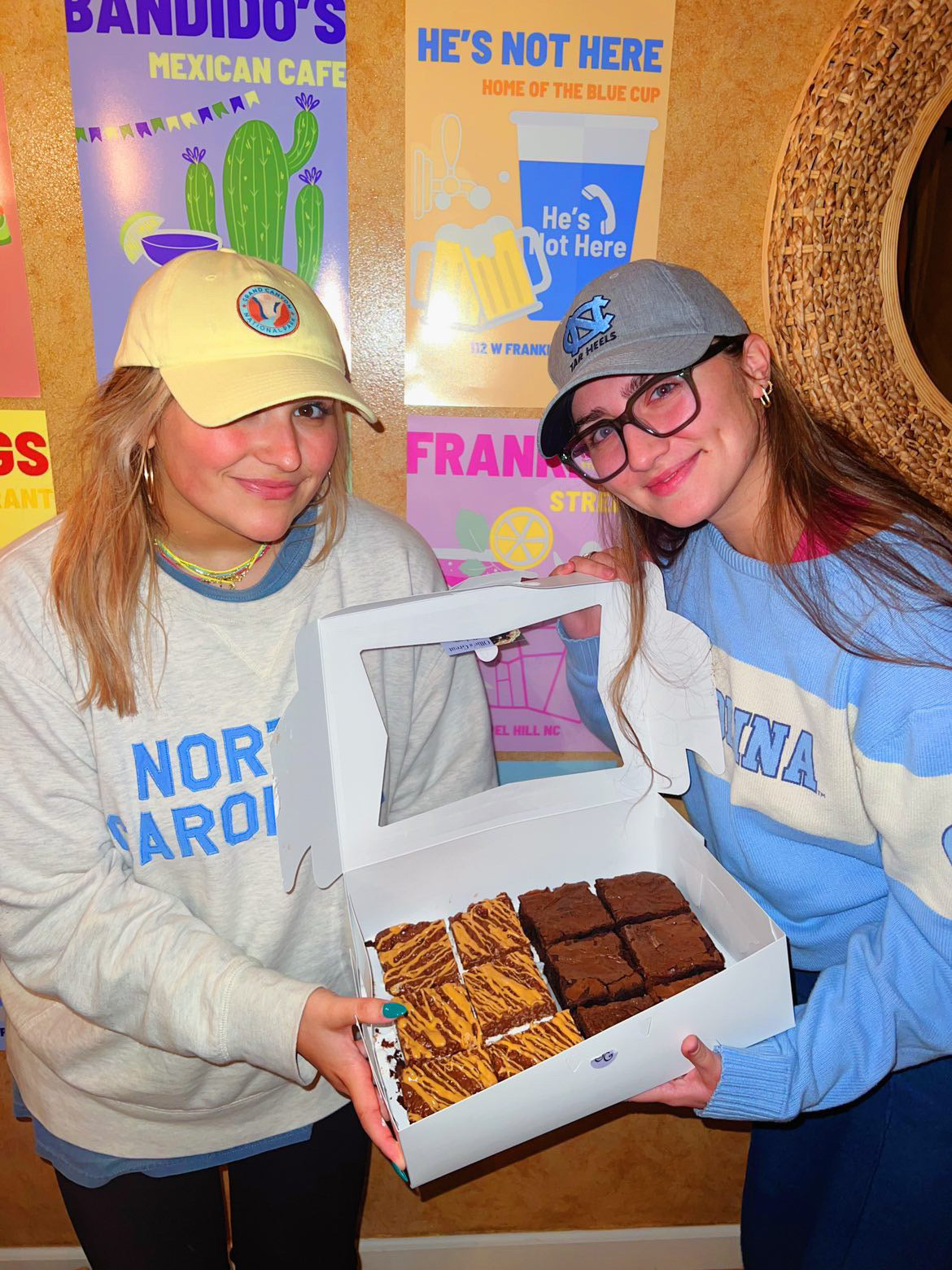 Two women holding a box of brownies in front of a colorful wall of local restaurant signs.