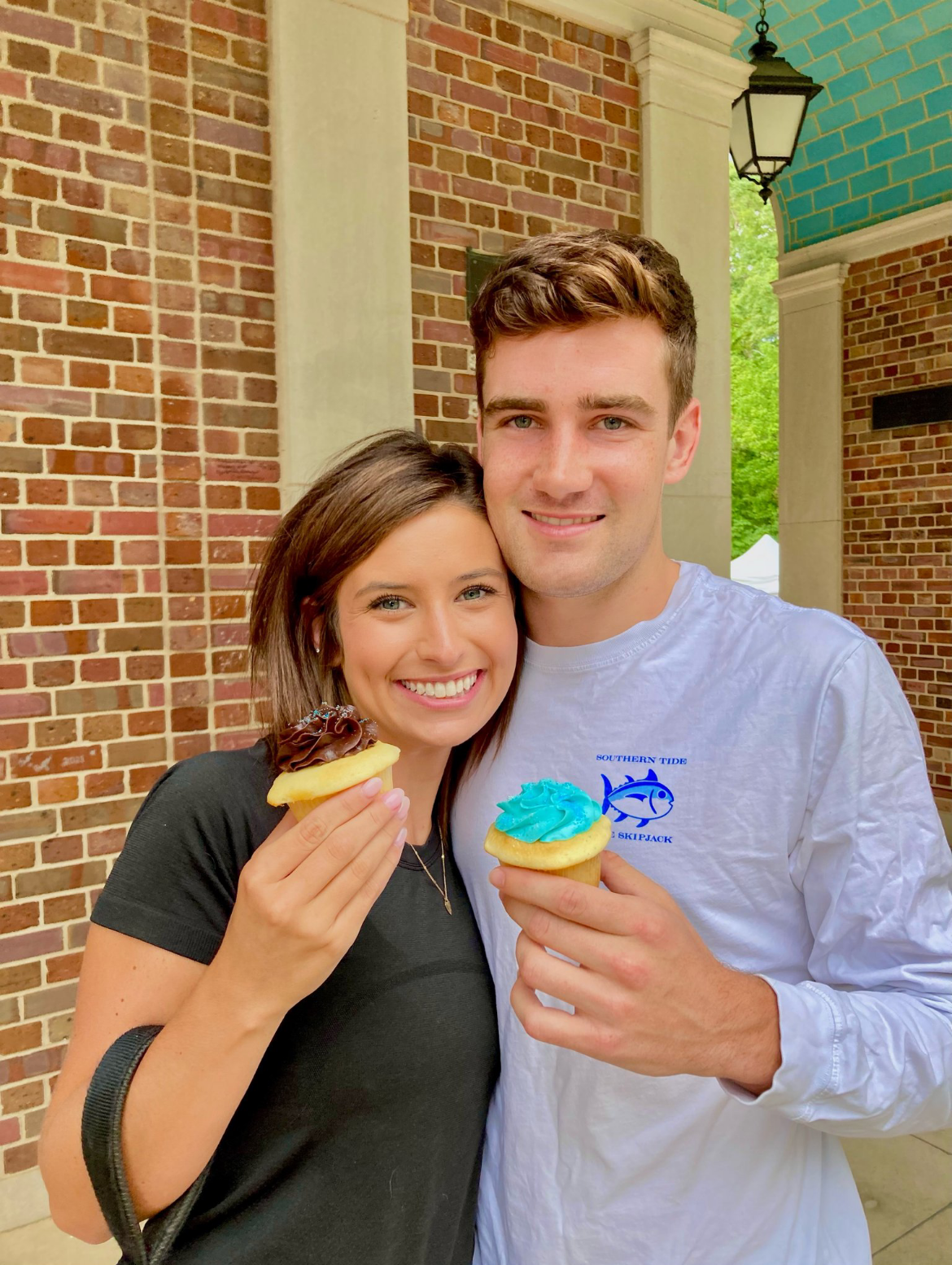 A man and woman holding cupcakes in front of a brick wall.