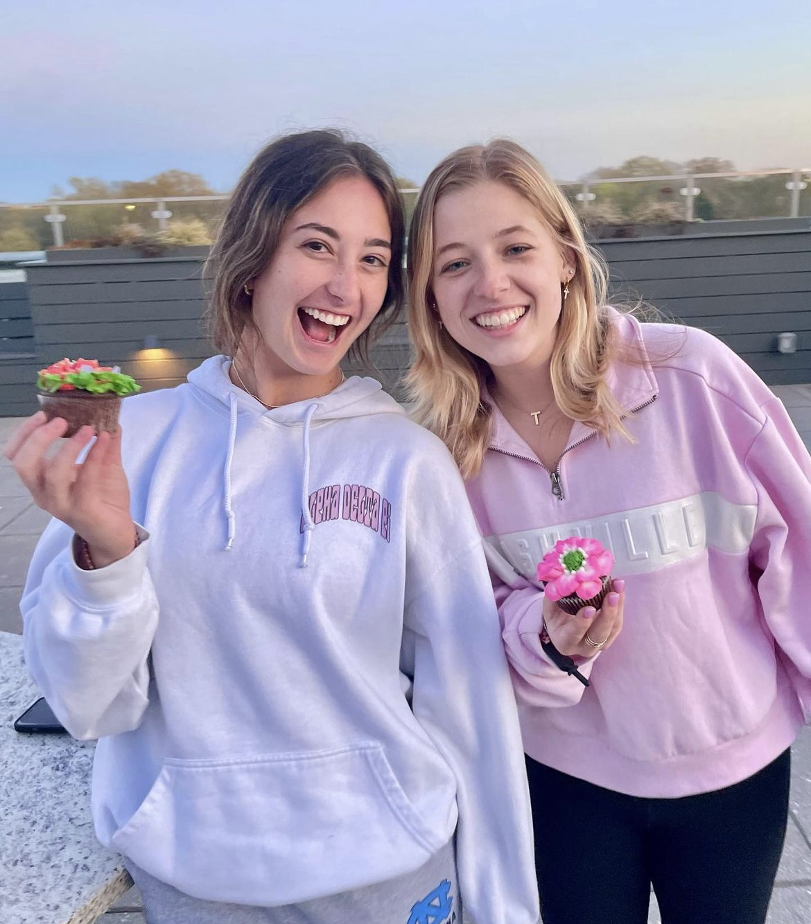Two people wearing hoodies holding floral cupcakes on a rooftop.