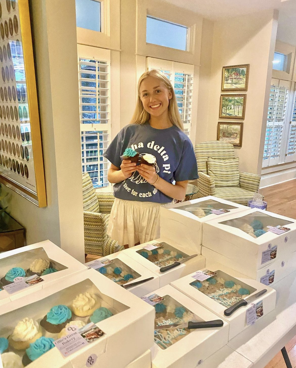UNC Student holding cupcakes behind a large number of catering boxes of cupcakes and cookie cakes.