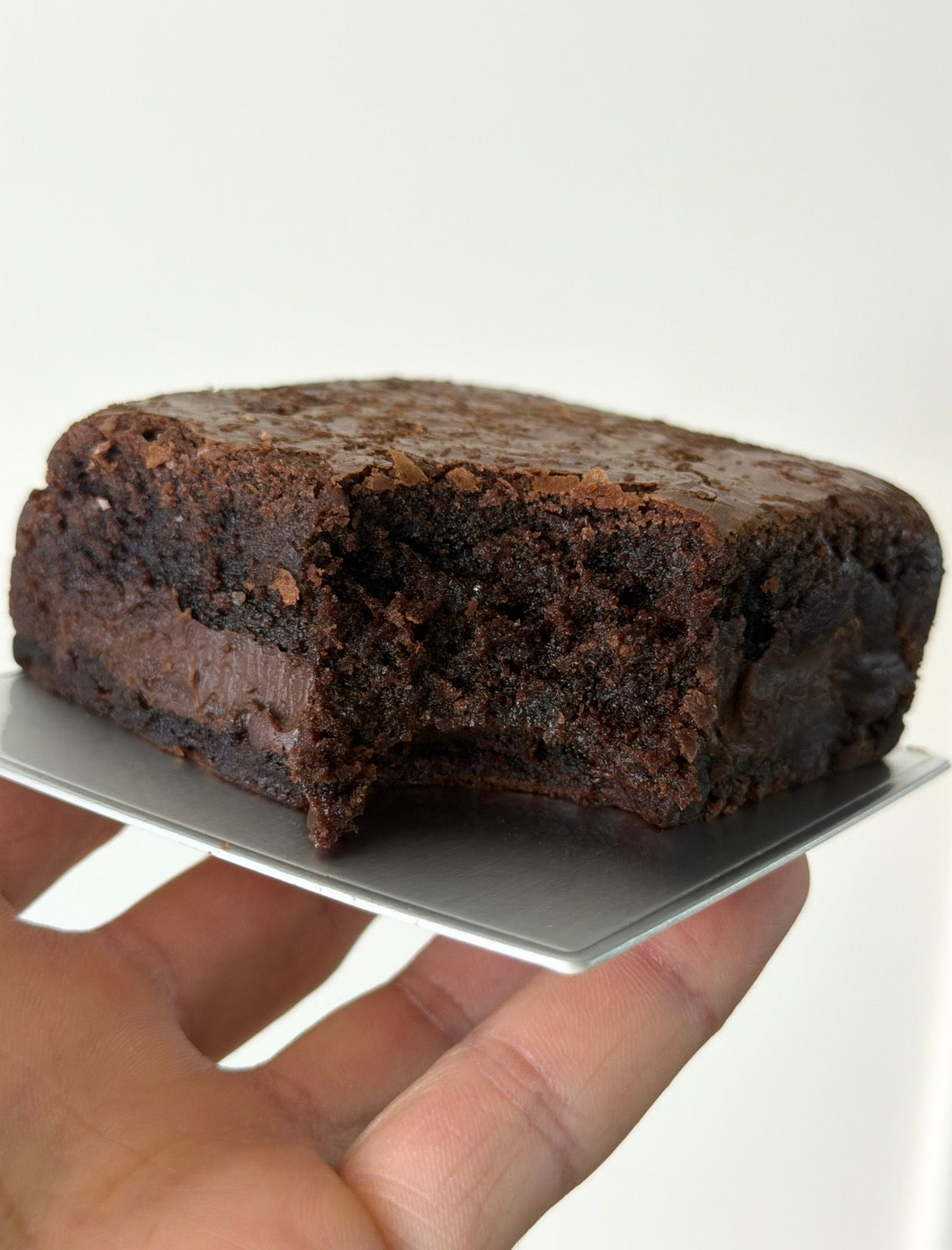A close-up of a hand holding a square silver tray with a rich, dark chocolate  fudge brownie, showing its moist, crumbly texture against a plain white background.
