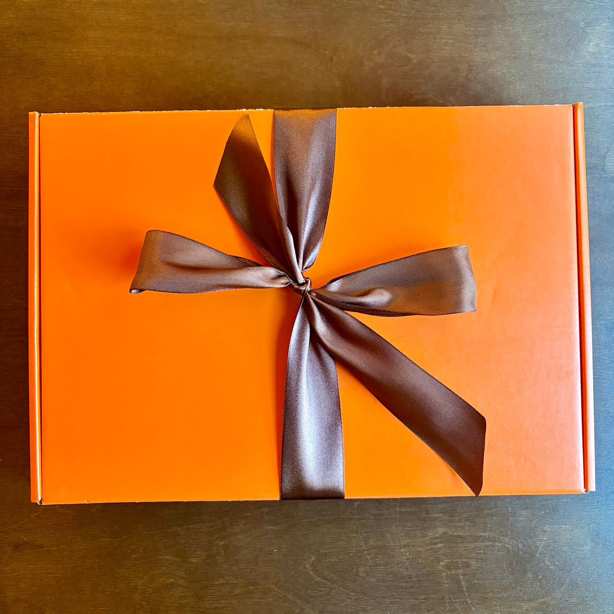 An orange rectangular gift box tied with a wide brown satin ribbon in a bow, photographed from above on a dark wood surface.