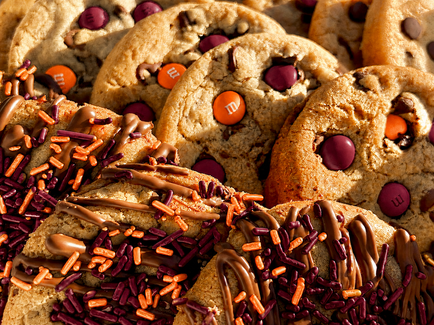 Close-up of maroon and orange M&M cookies and maroon and orange sprinkle cookies with chocolate drizzle.