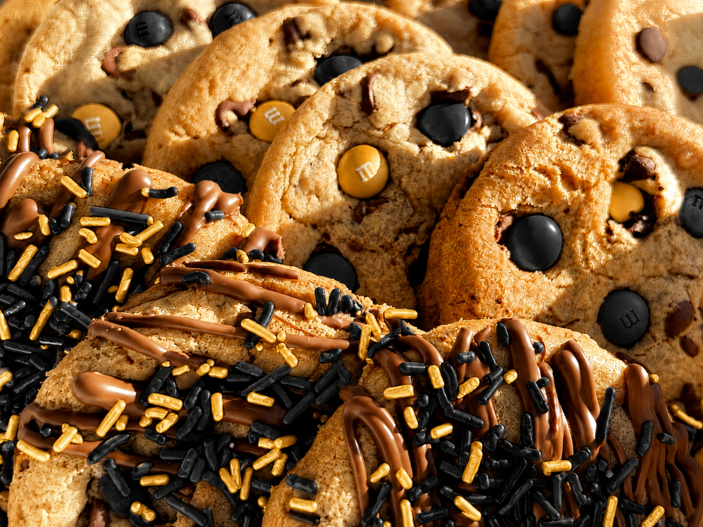 Close-up of black and gold M&M cookies and black and gold sprinkle cookies with chocolate drizzle.